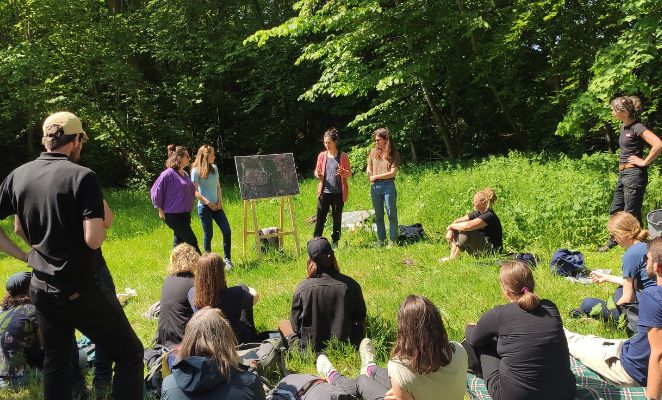 A group of people standing and sitting next to a blackboard, in a sunny glade in the forest.