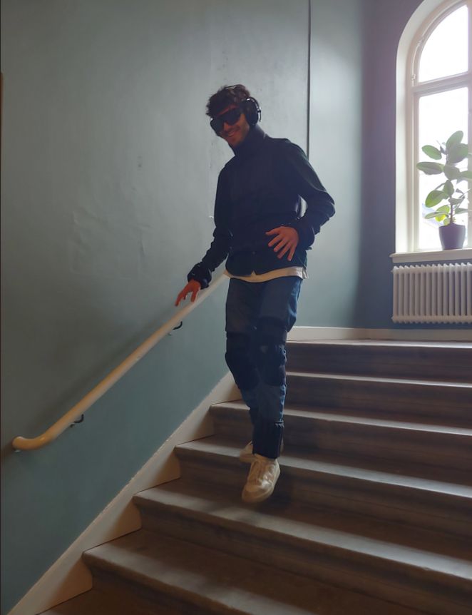 A young man goes down a flight of stairs. He's wearing various devices that simulate different kinds of disabilities. A weighted jacket that limits movement, and glasses that simulate visual impairments