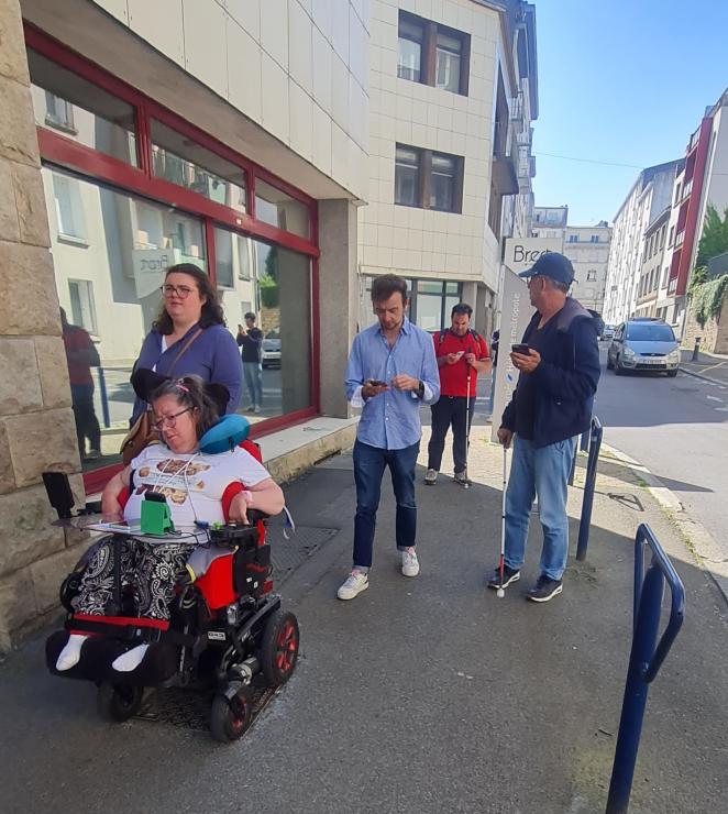 Five people are pictured on a sidewalk. One of them is using a motorised wheelchair. The others are holding their phones so they can analyse itineraries and report obstacles on the app