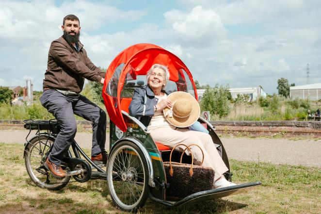 A person is shown being driven in an adapted vehicle: it's a rickshaw-like bike where the passenger is in the front and the driver is in the back