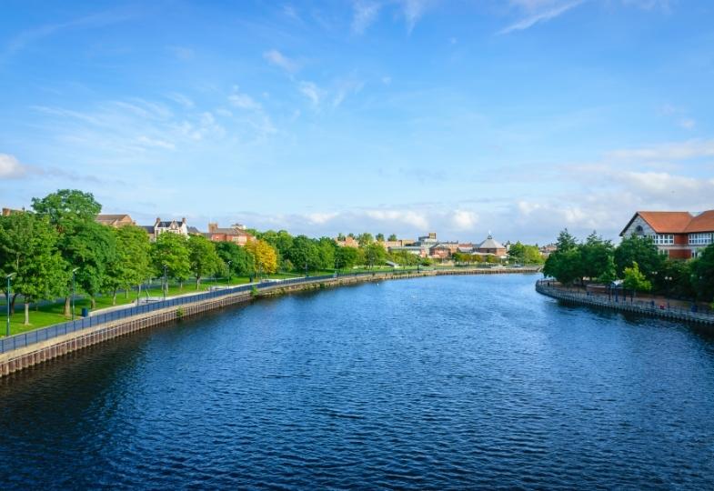 Tree-line river flowing past Stockton-on-Tees on a sunny day.