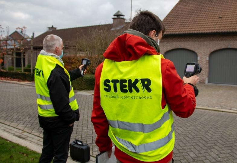 Two people wearing yellow safety vests standing in a street, holding devices that gauge heat loss from buildings.