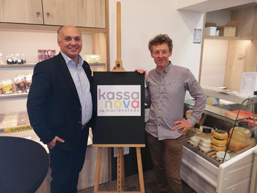 Two men in a shop, smiling at the camera and showing a blackboard with the text KASSA NOVA in coloured letters