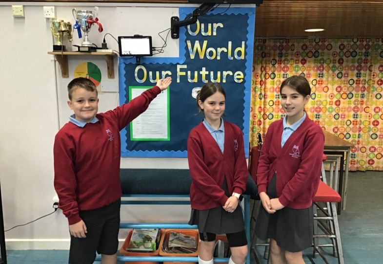 Three kids wearing school uniform in front of a large board with the text "Our world, our future"