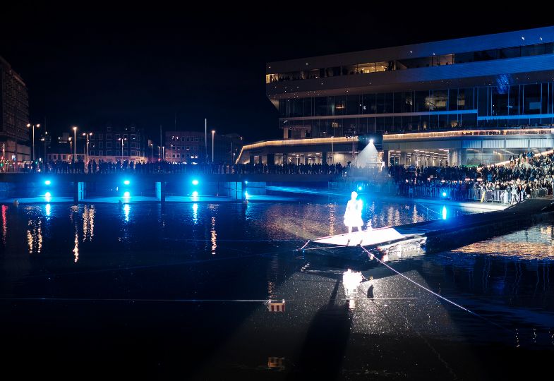 A light figure alone on water, while many people standing on the pier at night time