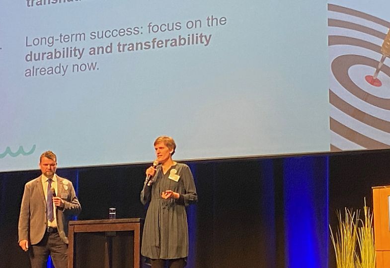 Two people in front of a large screen with the text "Long-term success: Focus on the durability and transferability already now