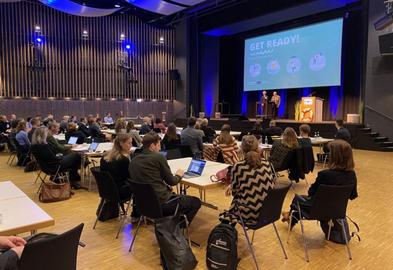 A plenary room full of applicants and two speakers on a stage in front of a large screen.