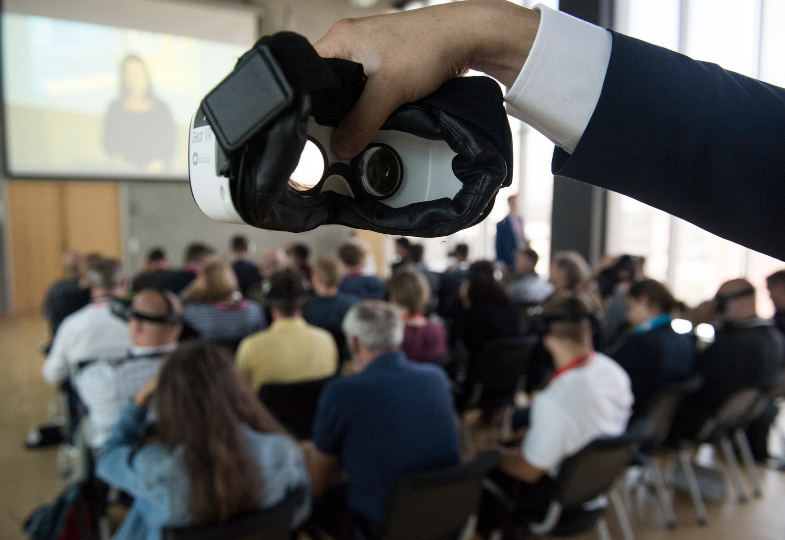 An arm holding VR glasses in front of a room ful of people looking at a screen