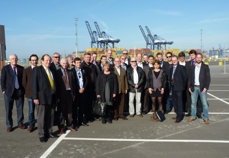 A group of people gathered in a harbour area with cranes against a clear sky.