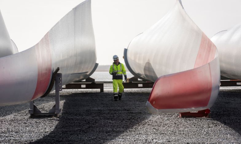 Worker with a helmet walking next to two wind turbine blades lying on the ground. 