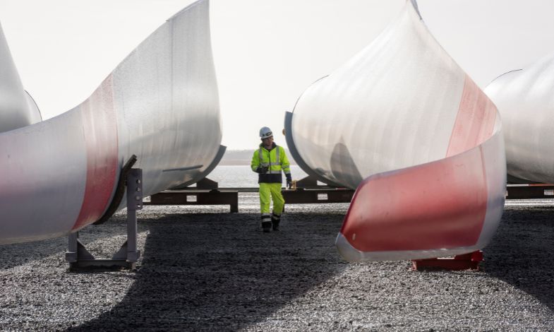 Worker with a helmet walking next to two wind turbine blades lying on the ground.