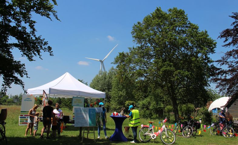 People gathering in a green landscape with white tents, trees, a blue sky and a wind turbine.