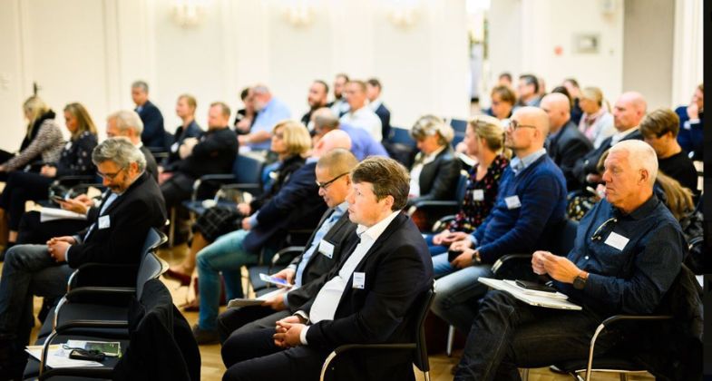 Audience at a conference, seated in rows in a light-filled room.