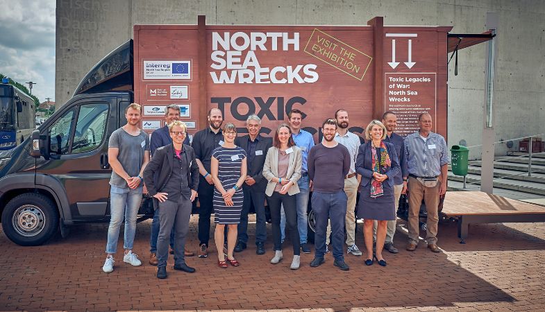A group of people in front of a collection of a vehicle titled North Sea Wrecks.