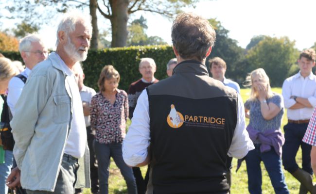 A group of people listening to a guide wearing a vest with a partridge embroidered on it.