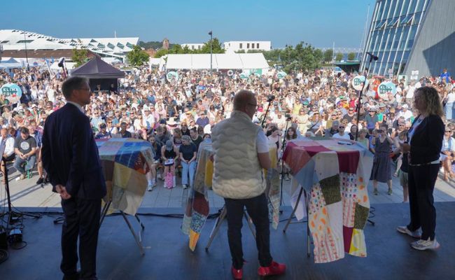 Three people on a stage overlooking a large crowd.