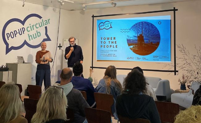 Two people giving a presentation in front of an audience. Two speech bubbles on the wall behind them say "Pop-up Circular Hub"