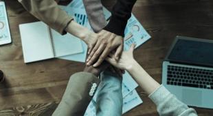 Four people's hands on top of each other across a table, with a laptop and some documents.