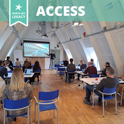 A small group of people in a white room with a wooden floor, lots of sunlight and sloping walls, listening to a presentation.