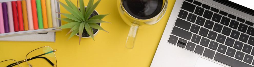 Coffee cup, laptop, plant, crayons, and glasses on a yellow table