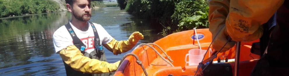 A man wearing big yellow gloves standing in the river next to a small orange boat