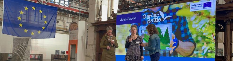 People at stage in front of a colourful screen showcasing a citizen climate challenge. A large EU flag is hanging down from the ceiling on the left.
