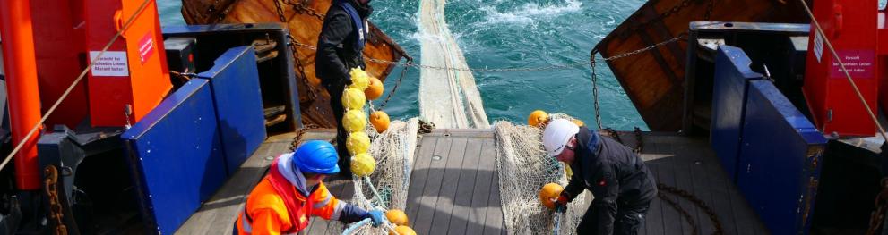 Three people wearing safety helmets and protective clothing, standing on the deck of a boat working on a net trailing behind the boat.