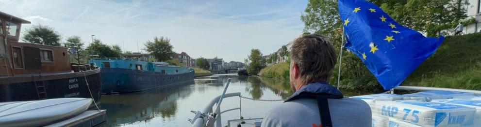 An man steering a barge with a waving EU flag across a river.