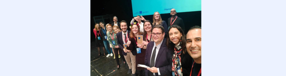 A group of people is on the stage of the InterregSlam event. In the centre, Esen, who represented SMALL at the event, holds the award in her hands. Everyone is smiling and celebrating the win.