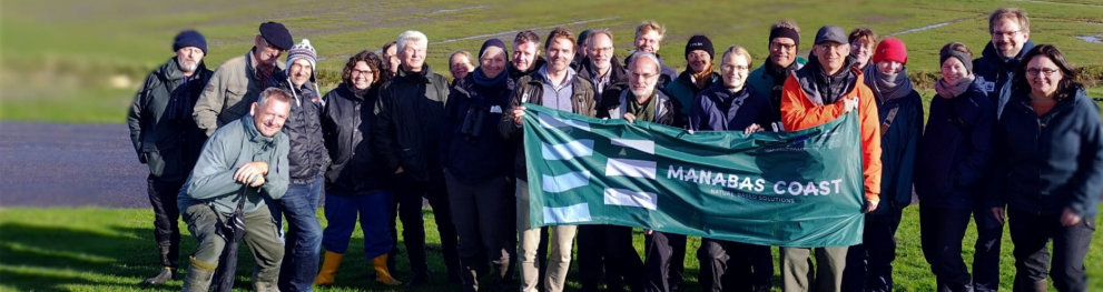 A group of people standing in a green field next to the sea, holding a green banner saying MANABAS COAST.