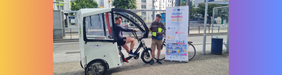 Nicholas Hastings sits on the rickshaw he drove for the project discussed. A roll-up poster of the SMALL project is visible on the right