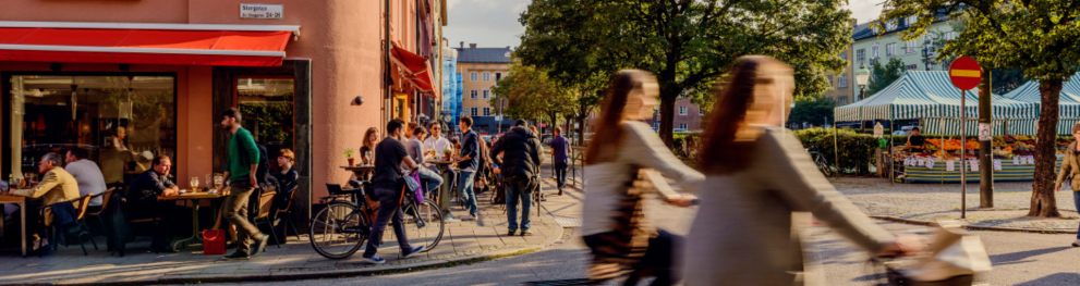 Two people biking past a busy café in Malmö on a sunny day. Credits to Malmö Stad.