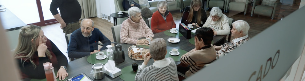 A group of older adults and care staff sit around a table in a care facility, having coffee and conversation, while a caregiver stands nearby observing the interaction.